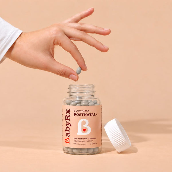 A hand reaches down to pick up a single softgel from a clear bottle labeled BabyRx Complete Postnatal, with the gray pill placed beside the bottle on a warm beige background.